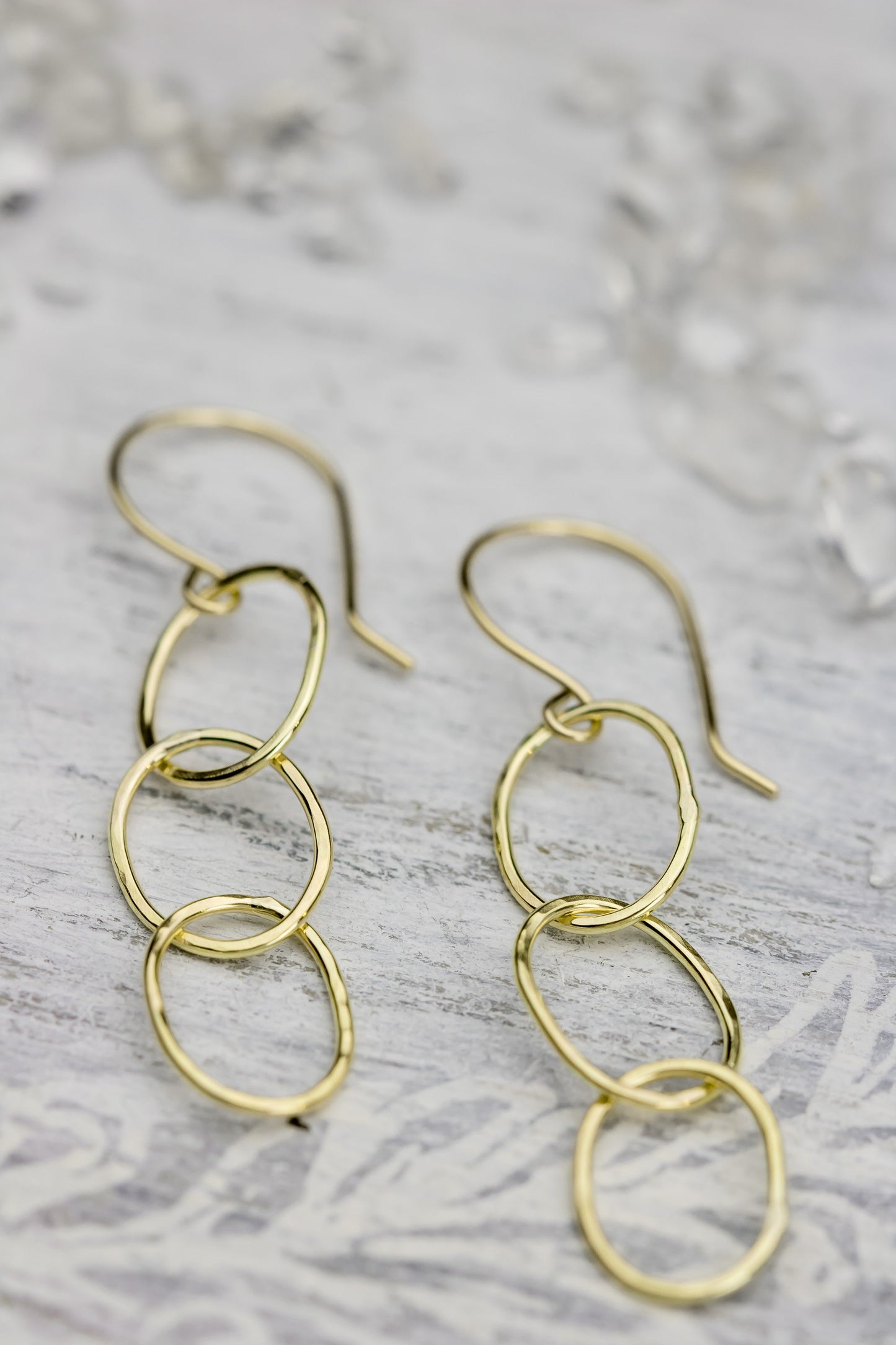 Two 18K yellow gold hammered triple hoop earrings placed on a white textured surface with a faint floral pattern, highlighting their design.