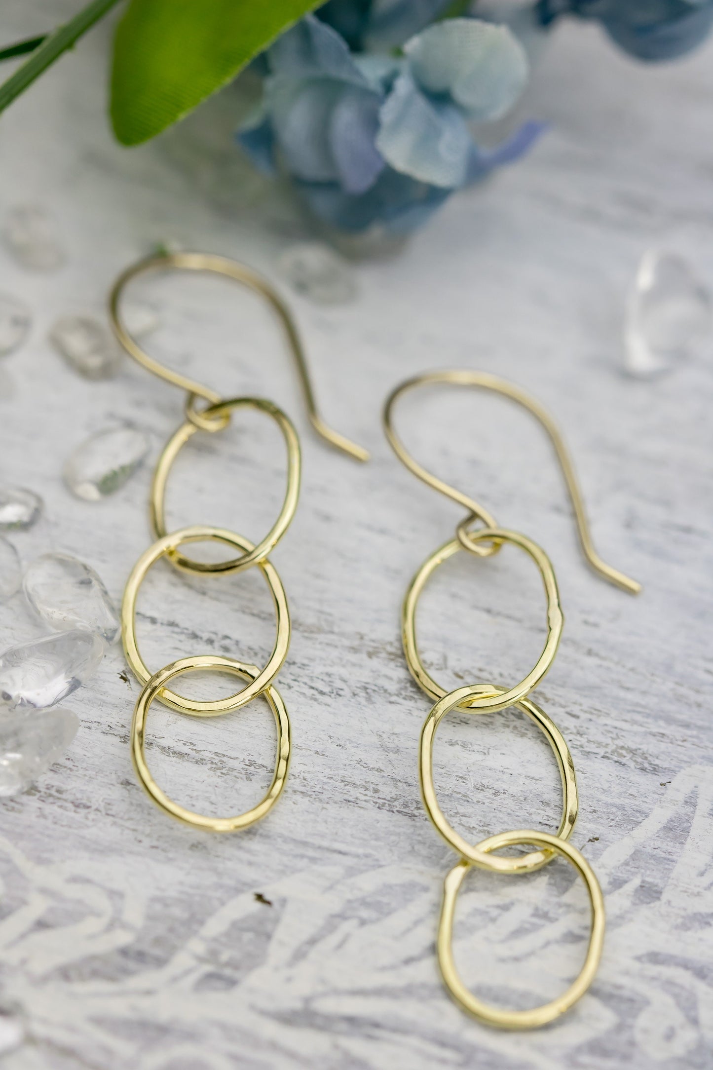 Two 18K yellow gold hammered triple hoop earrings placed on a white textured surface with a faint floral pattern, highlighting their design.