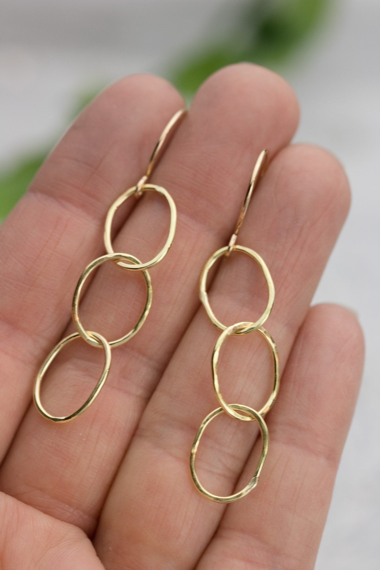 A hand holding a pair of 18K yellow gold hammered triple hoop earrings, displaying their three linked hoops and textured surface.