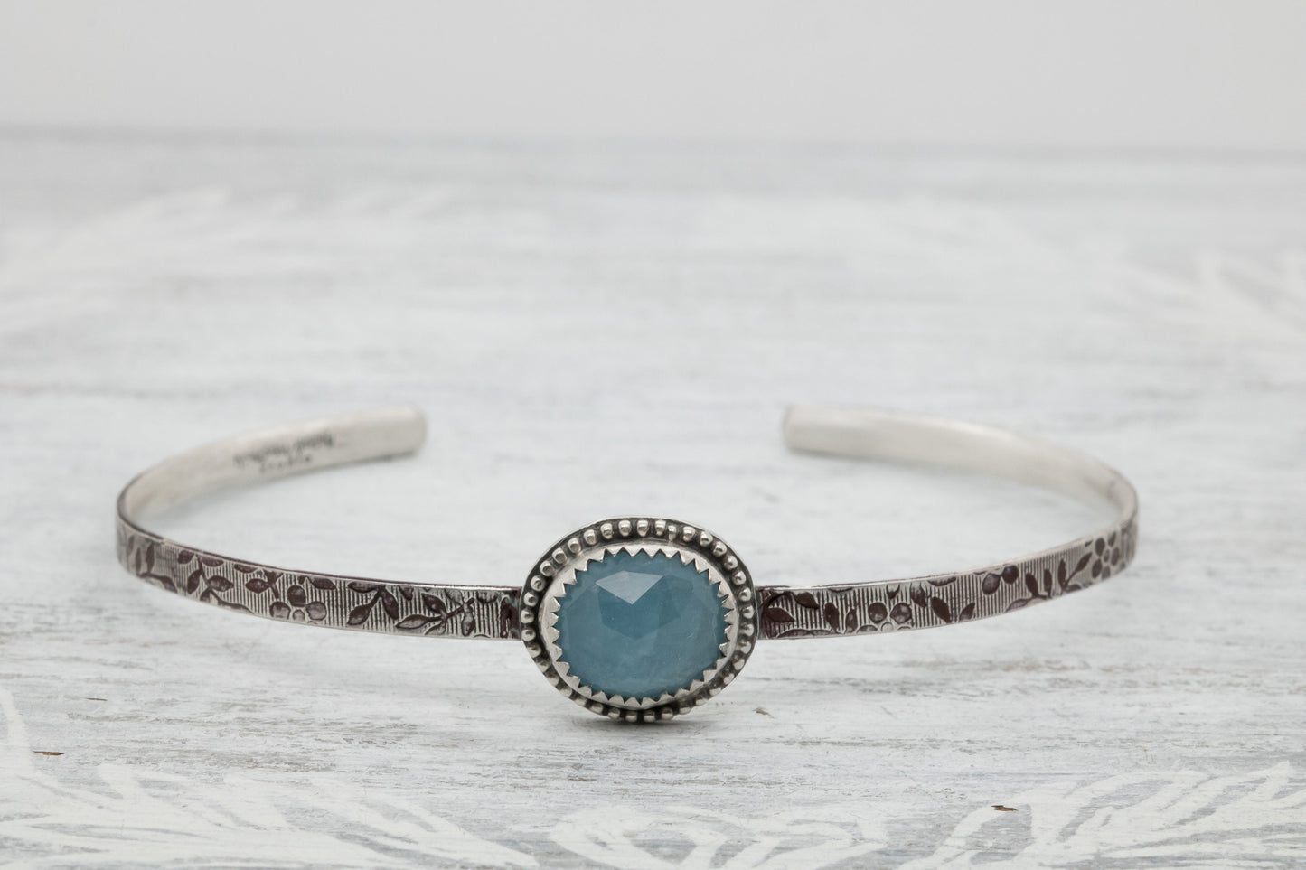 Silver bracelet with floral engravings, featuring a oval blue aquamarine on a light wood surface with scattered white baby breath flower.