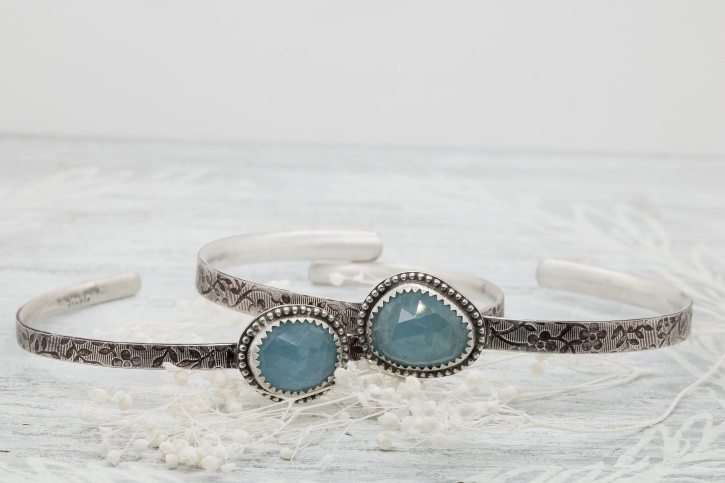 Silver bracelet with floral engravings, featuring a oval blue aquamarine on a light wood surface with scattered white baby breath flower.
