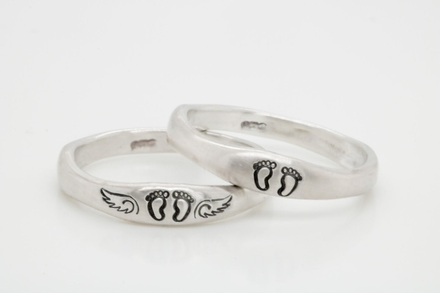 Two sterling silver rings with hand-stamped baby feet and wings, placed on a white wooden surface with delicate white flowers, showcasing their 4mm width and stackable design.