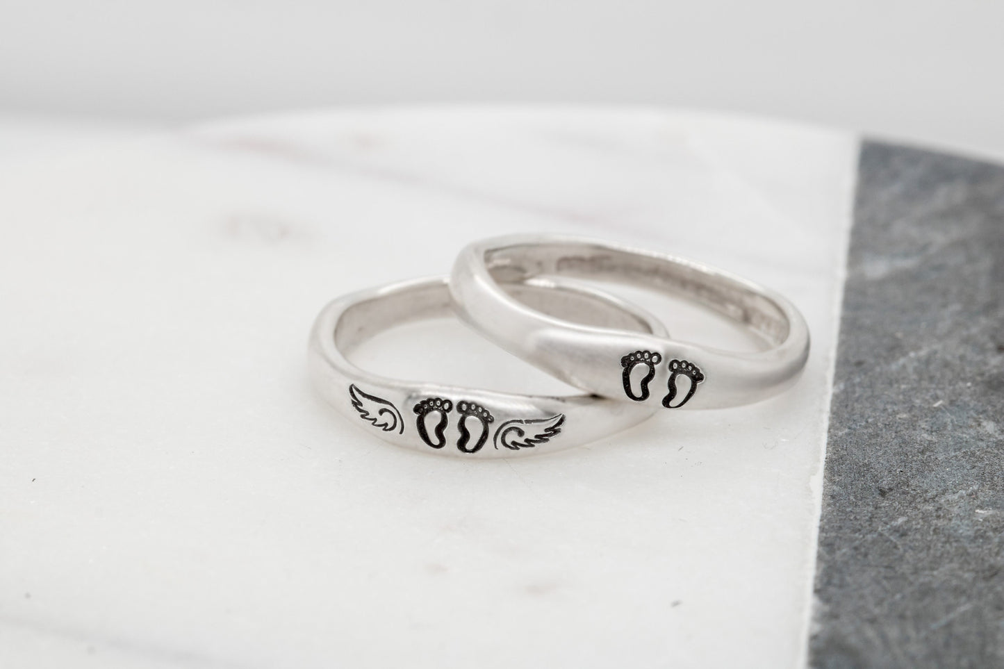 Two sterling silver rings with hand-stamped baby feet and wings, placed on a white wooden surface with delicate white flowers, showcasing their 4mm width and stackable design.