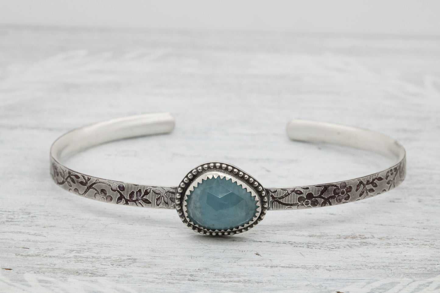 Silver bracelet with floral engravings, featuring a oval blue aquamarine on a light wood surface with scattered white baby breath flower.