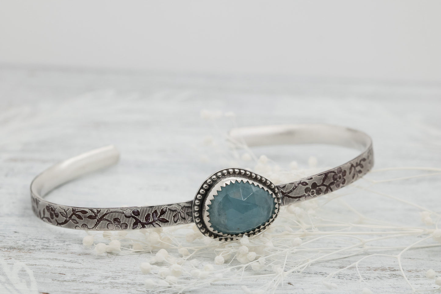 Silver bracelet with floral engravings, featuring a oval blue aquamarine on a light wood surface with scattered white baby breath flower.