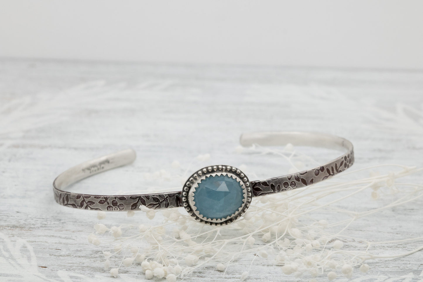 Silver bracelet with floral engravings, featuring a oval blue aquamarine on a light wood surface with scattered white baby breath flower.