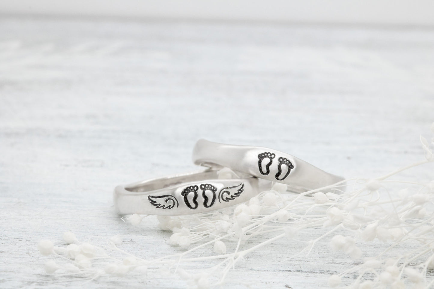 Two sterling silver rings with hand-stamped baby feet and wings, placed on a white wooden surface with delicate white flowers, showcasing their 4mm width and stackable design.