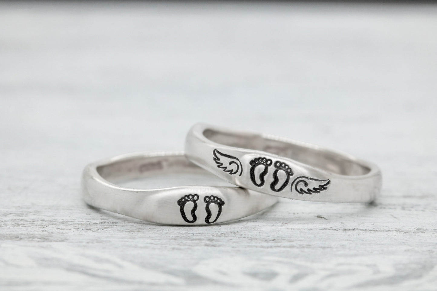 Two sterling silver rings with hand-stamped baby feet and wings, placed on a white wooden surface with delicate white flowers, showcasing their 4mm width and stackable design.