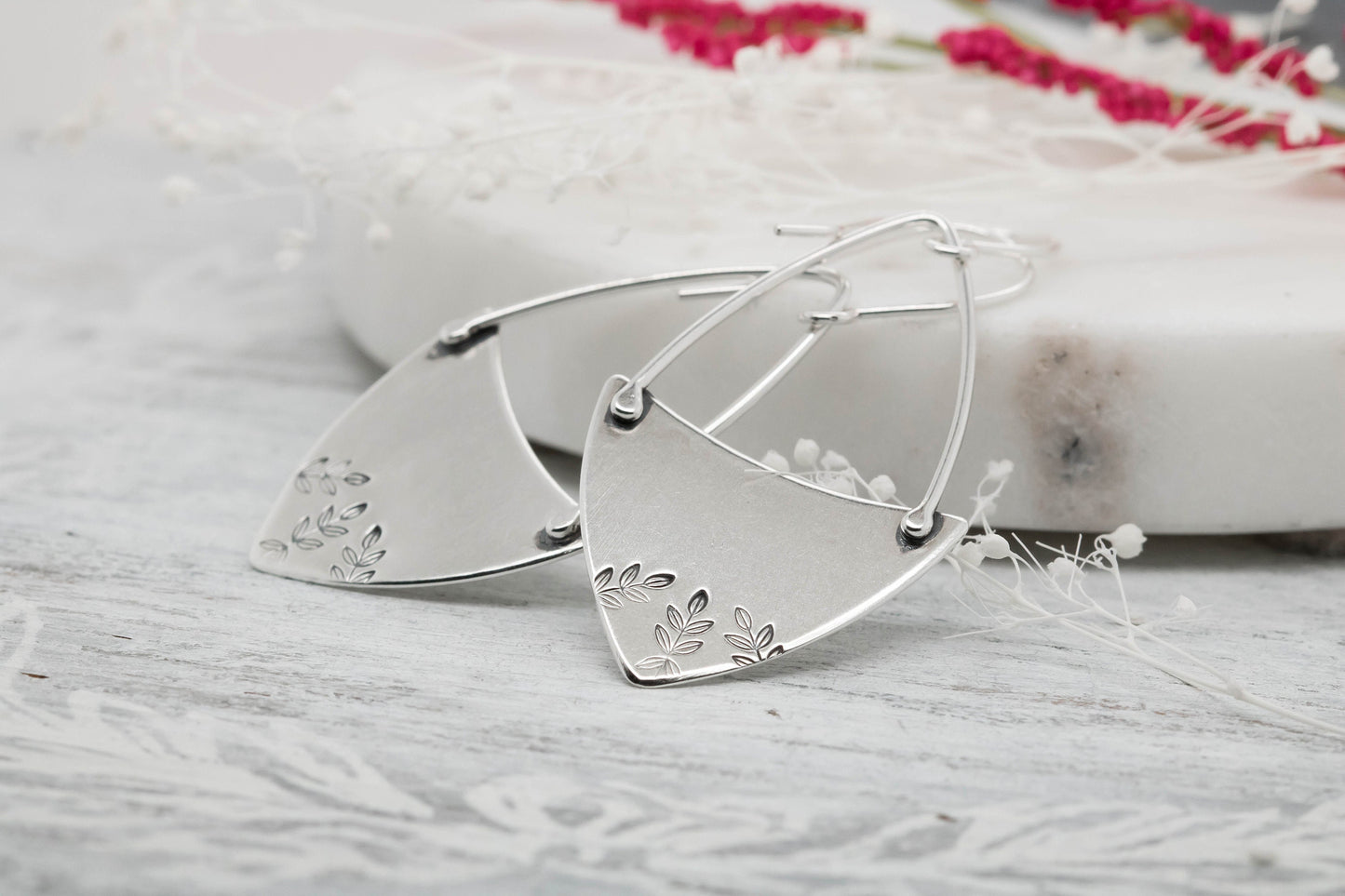 A pair of sterling silver hoop earrings with a floral fern design, displayed against a natural backdrop.