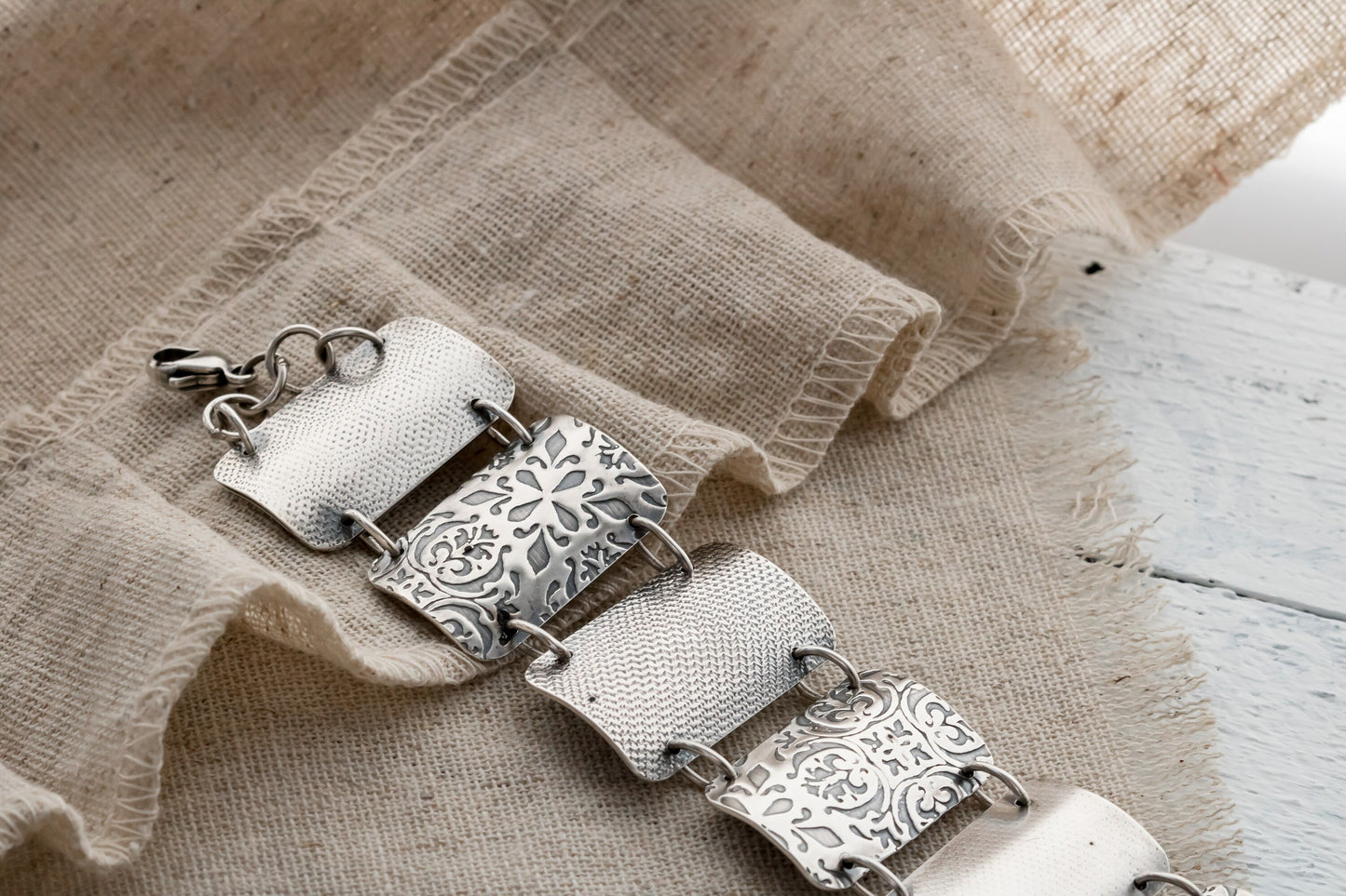 A close-up image of a silver bracelet with intricate designs, resting on a textured beige fabric. The bracelet features four wide, curved segments, each adorned with detailed floral and geometric engravings. The segments are connected by small metal loops, and a clasp is visible on the left side.