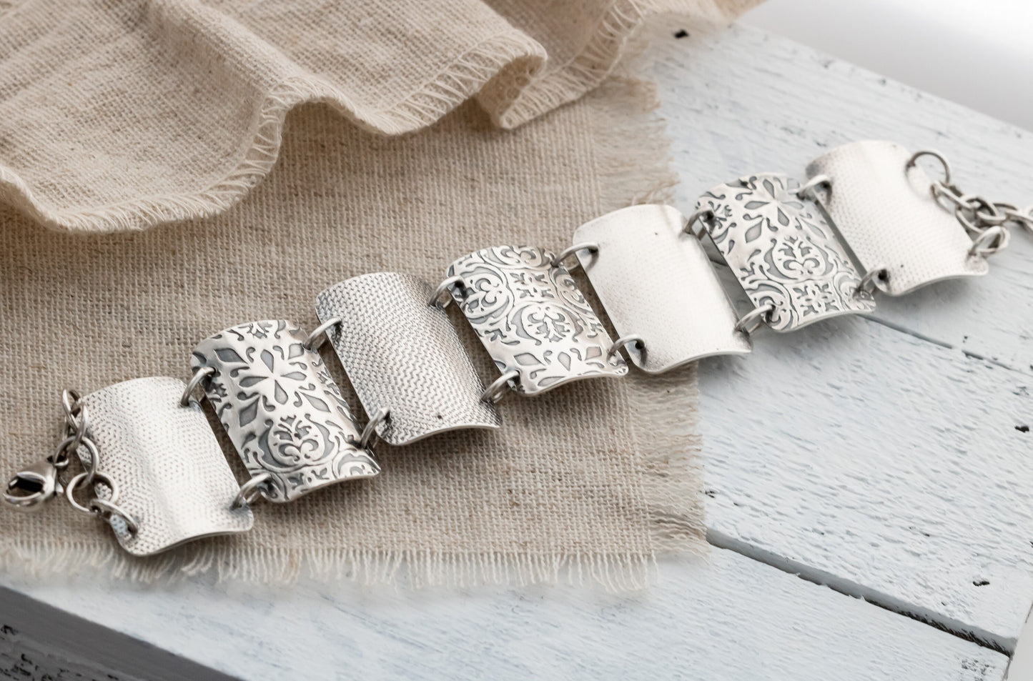 A close-up image of a silver bracelet with intricate designs, resting on a textured beige fabric. The bracelet features four wide, curved segments, each adorned with detailed floral and geometric engravings. The segments are connected by small metal loops, and a clasp is visible on the left side.