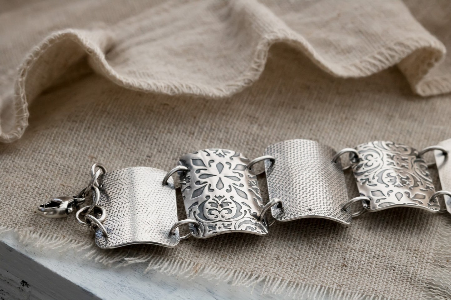 A close-up image of a silver bracelet with intricate designs, resting on a textured beige fabric. The bracelet features four wide, curved segments, each adorned with detailed floral and geometric engravings. The segments are connected by small metal loops, and a clasp is visible on the left side.