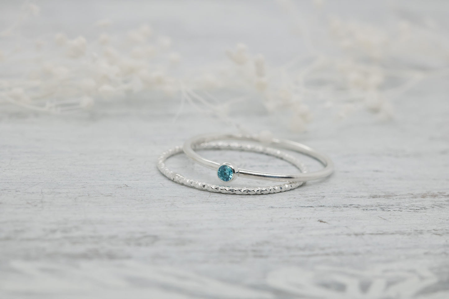 Dainty sterling silver stacking rings with a blue cubic zirconia birthstone, displayed on a textured white surface with dried flowers in the background.