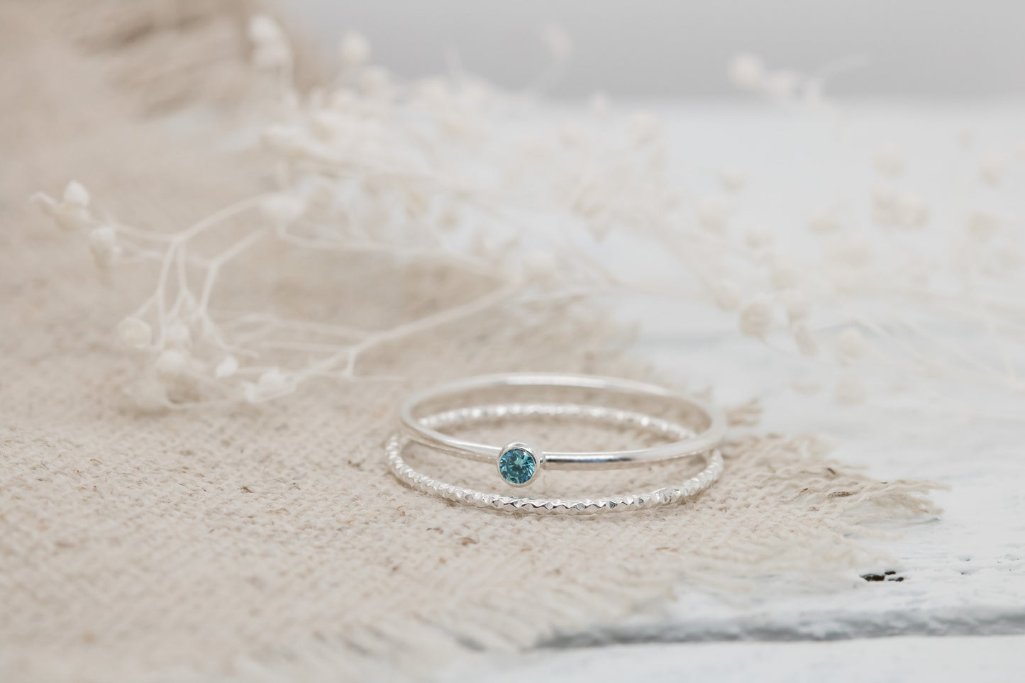 Dainty sterling silver stacking rings with a blue cubic zirconia birthstone, displayed on a textured white surface with dried flowers in the background.
