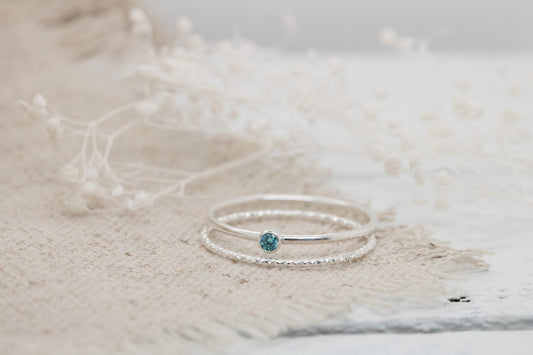 Dainty sterling silver stacking rings with a blue cubic zirconia birthstone, displayed on a textured white surface with dried flowers in the background.