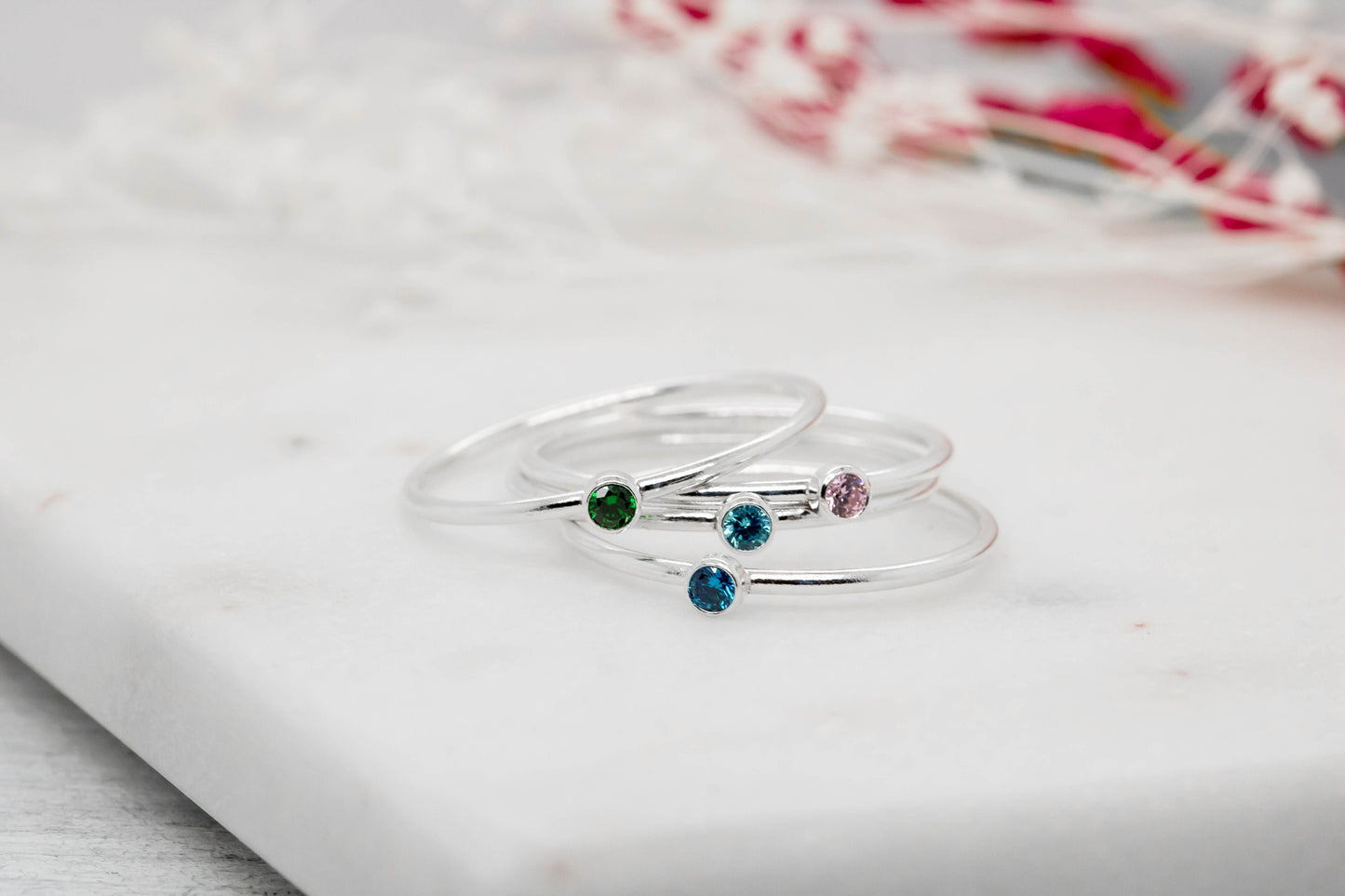 Dainty sterling silver stacking rings with a blue cubic zirconia birthstone, displayed on a textured white surface with dried flowers in the background.