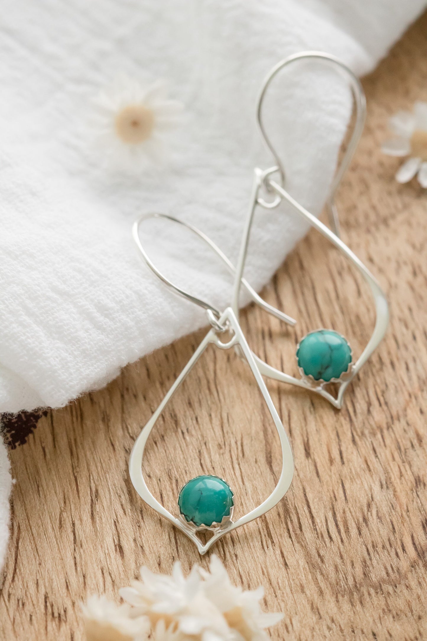 Pair of silver drop earrings with turquoise gemstones, resting on a white fabric with dried flowers on a wooden Surface.