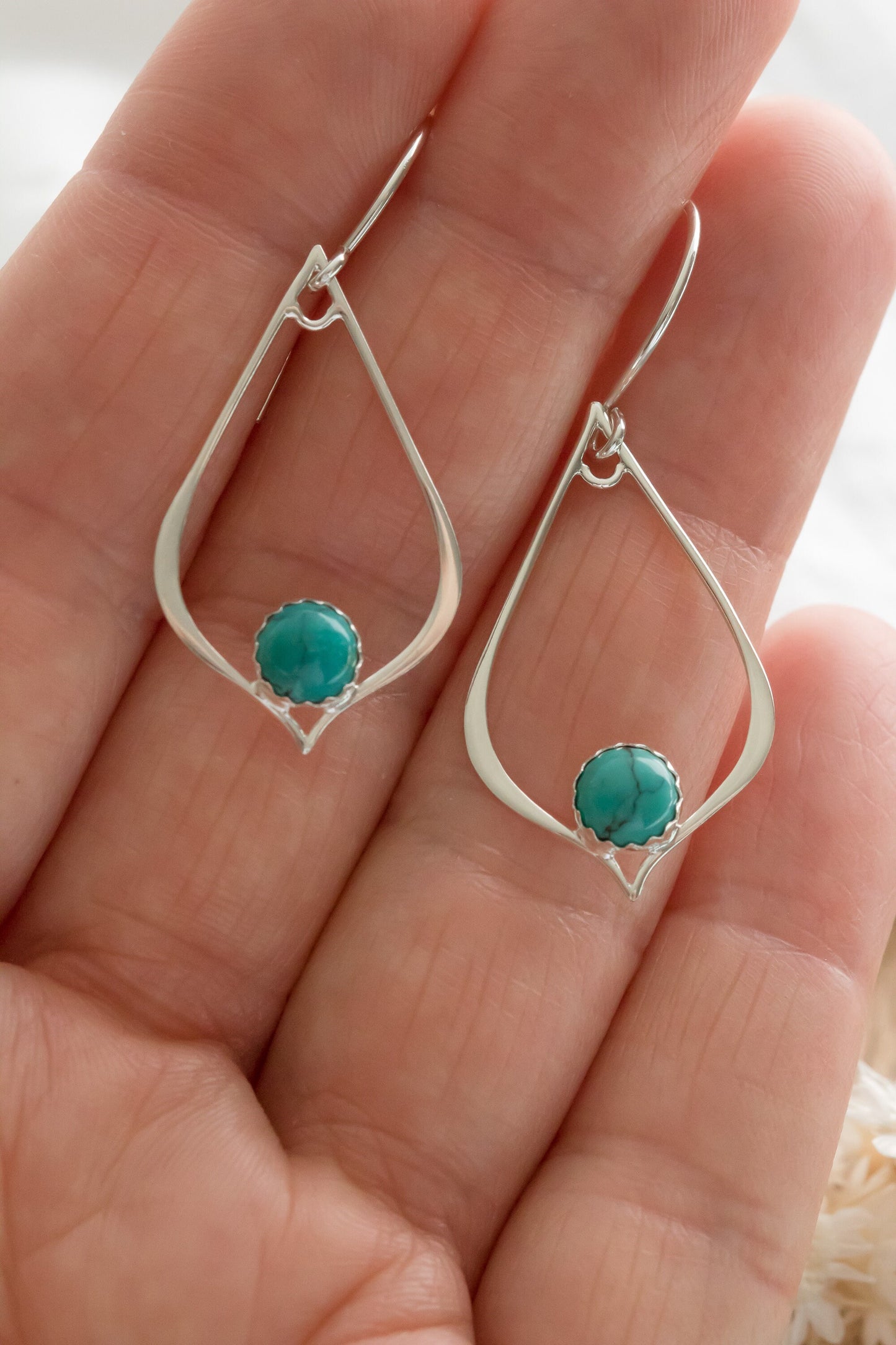 Close-up of a hand holding two silver teardrop-shaped earrings, each featuring a turquoise gemstone set in a detailed bezel.