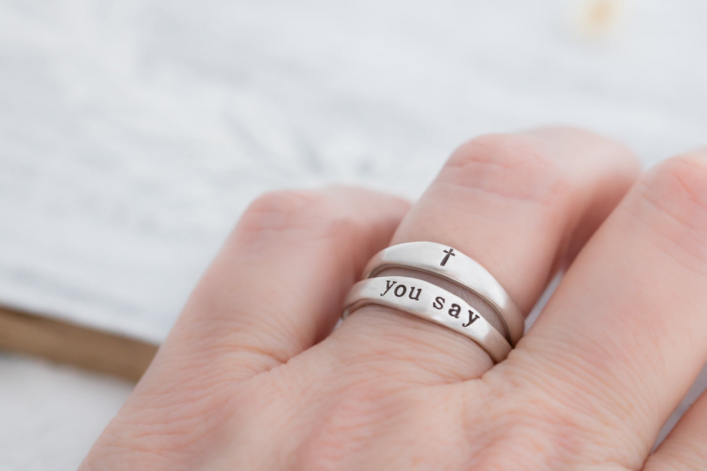 A hand wearing two sterling silver stackable rings, one engraved with "YOU SAY" and a cross symbol, placed over an open book with a soft-focus background.