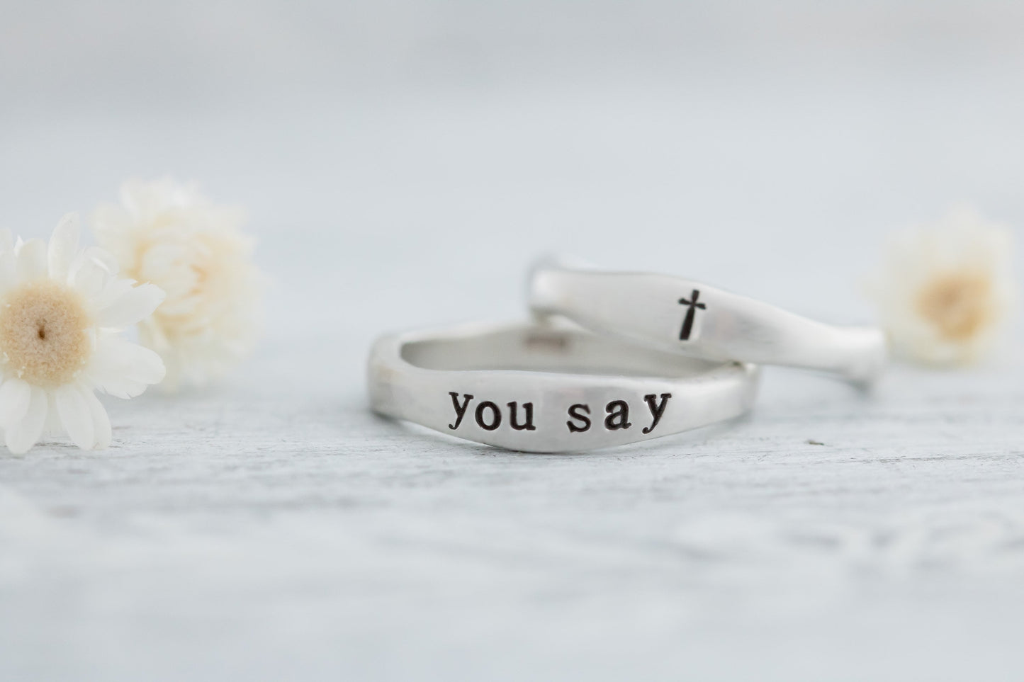 Two sterling silver rings, one engraved with "YOU SAY" and a cross, displayed on a white wooden surface with dried white flowers in the background.