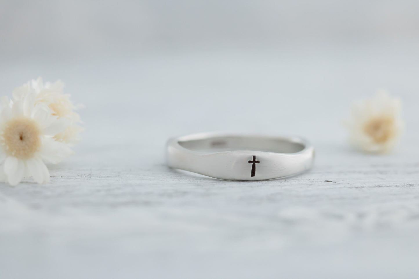 Sterling silver ring engraved with a cross, displayed on a white wooden surface with dried white flowers in the background.
