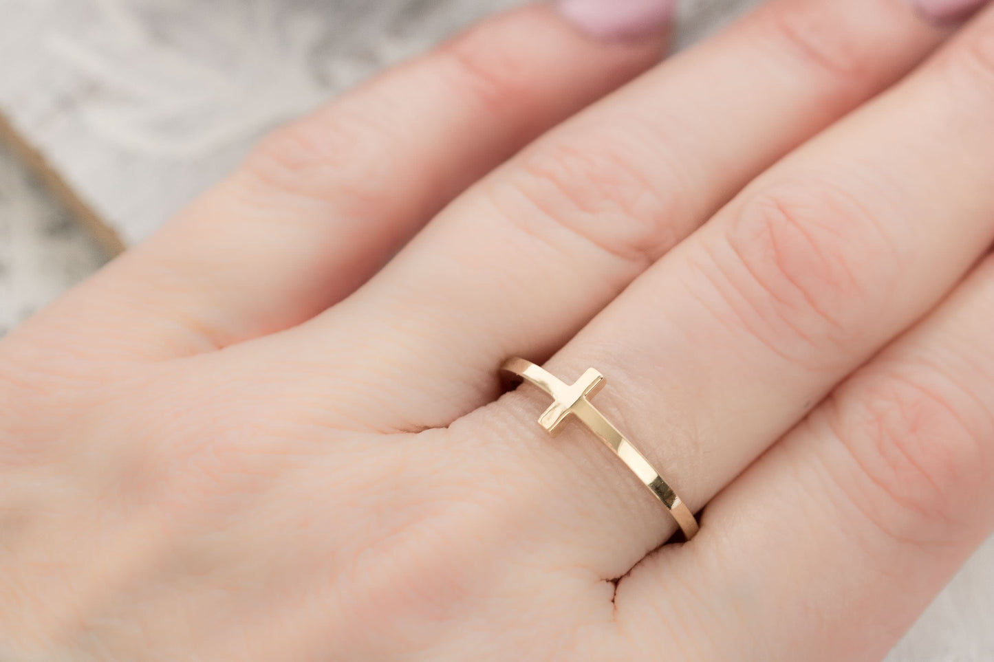 Alt Text 1: A close-up of a solid 14k gold sideways cross ring resting on delicate white dried leaves against a light wooden background, showcasing its elegant design.