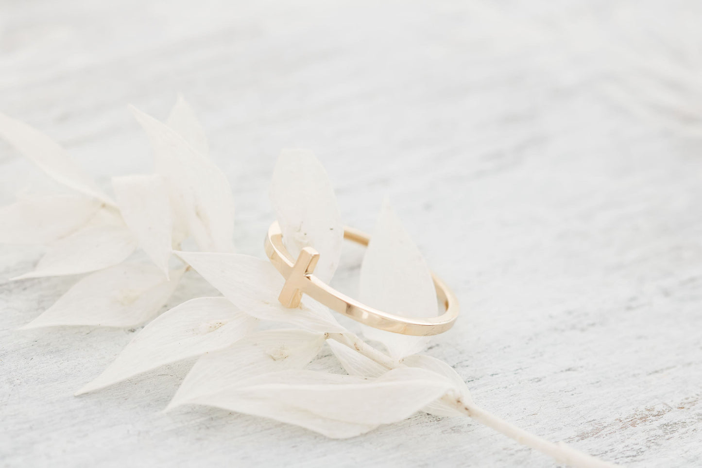 A close-up of a solid 14k gold sideways cross ring resting on delicate white dried leaves against a light wooden background, showcasing its elegant design.
