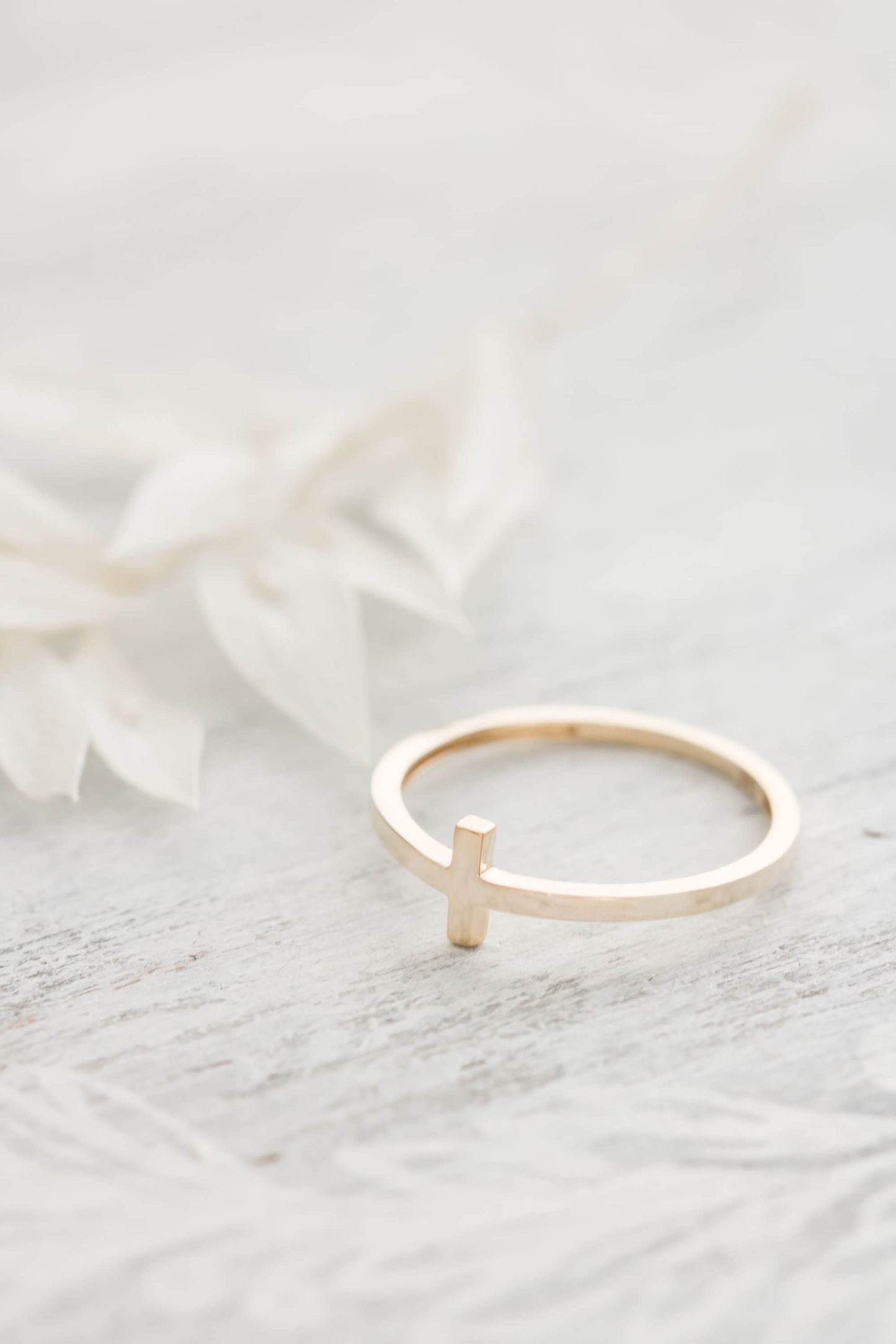 Alt Text 1: A close-up of a solid 14k gold sideways cross ring resting on delicate white dried leaves against a light wooden background, showcasing its elegant design.