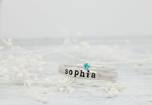 Sterling silver name ring with "Sophia" and blue birthstone on white floral background.