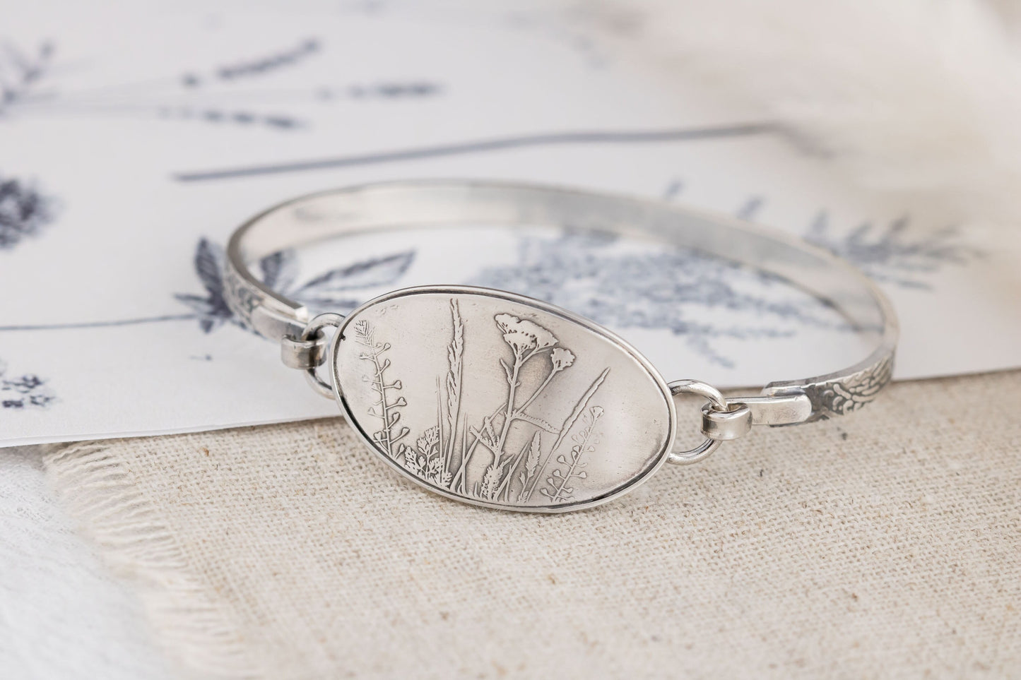 Textured sterling silver bangle bracelet with a floral design, displayed on a piece of paper.