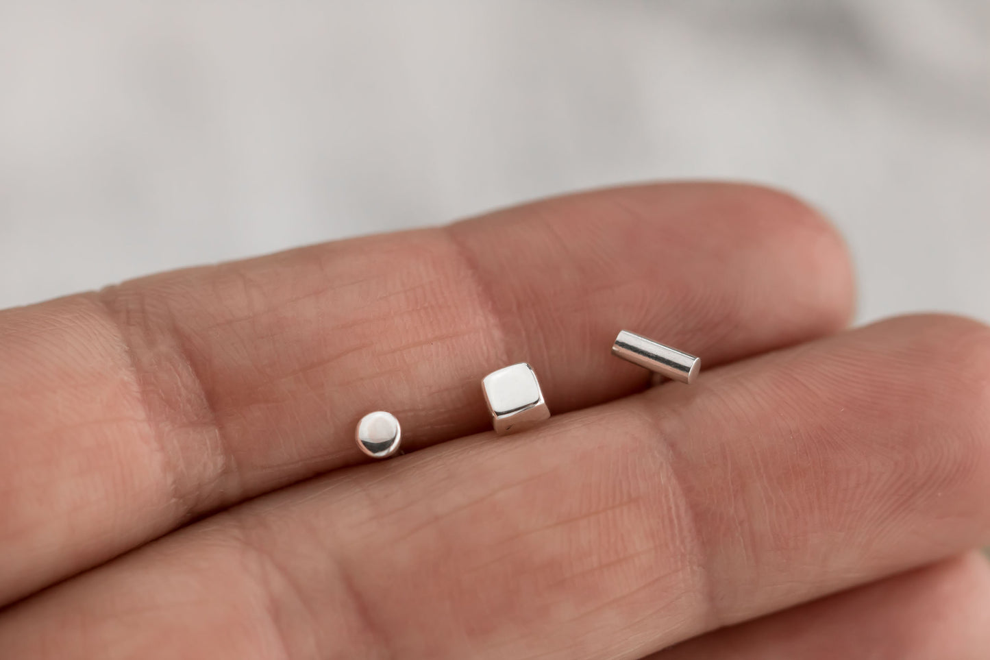 Close-up of a hand holding a set of minimalist sterling silver stud earrings, including dot, square, and bar designs.