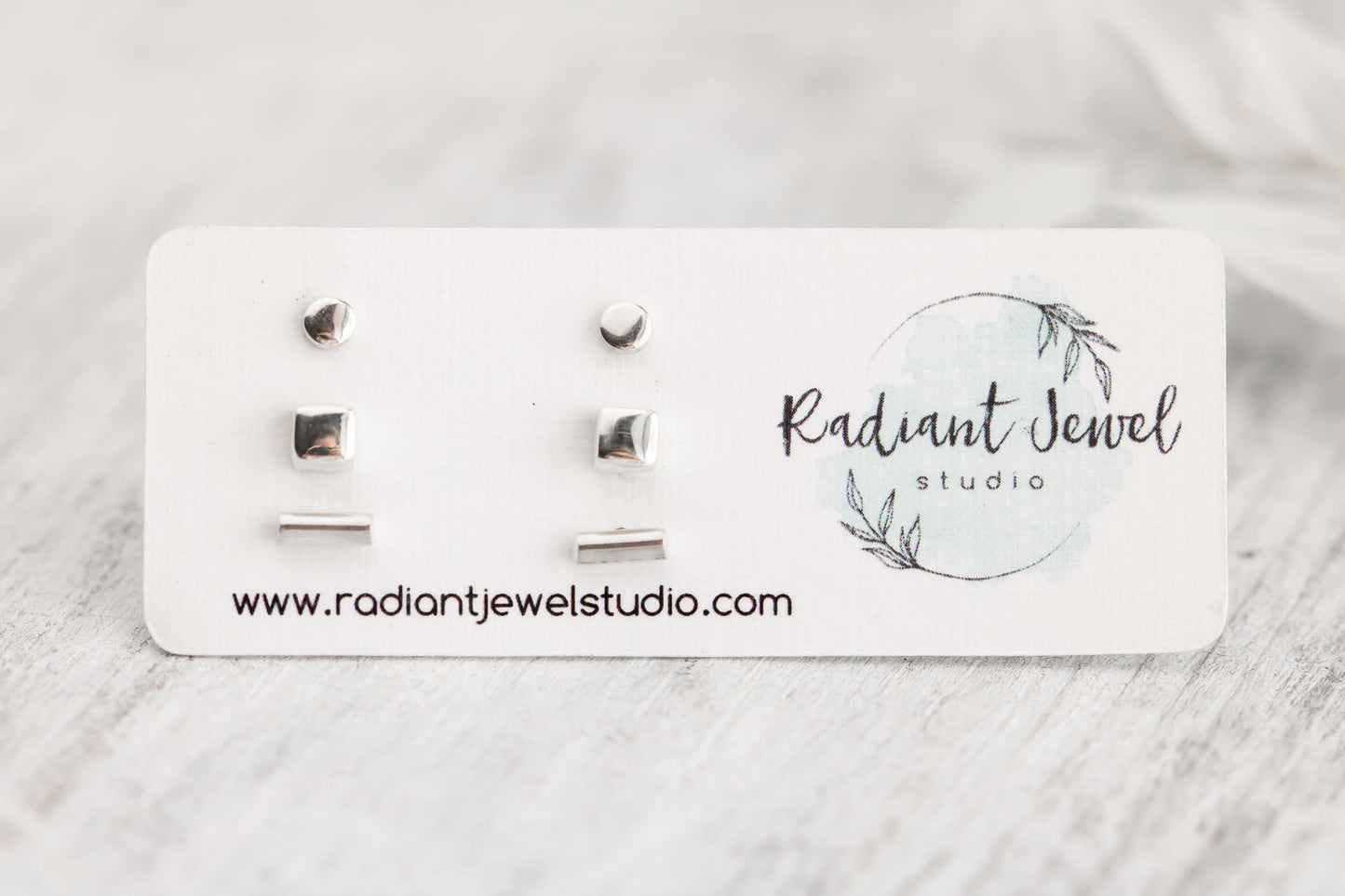 A set of minimalist sterling silver stud earrings presented on a white card with the Radiant Jewel Studio logo, featuring three different shapes: a dot, a square, and a bar.