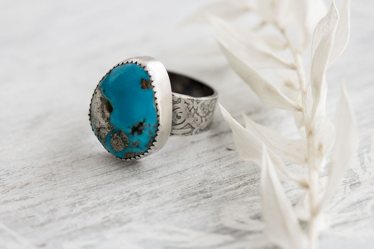 A sterling silver ring with a hand-cut Campitos turquoise stone featuring pyrite speckles, placed on a white wooden surface with dried white leaves in the background.