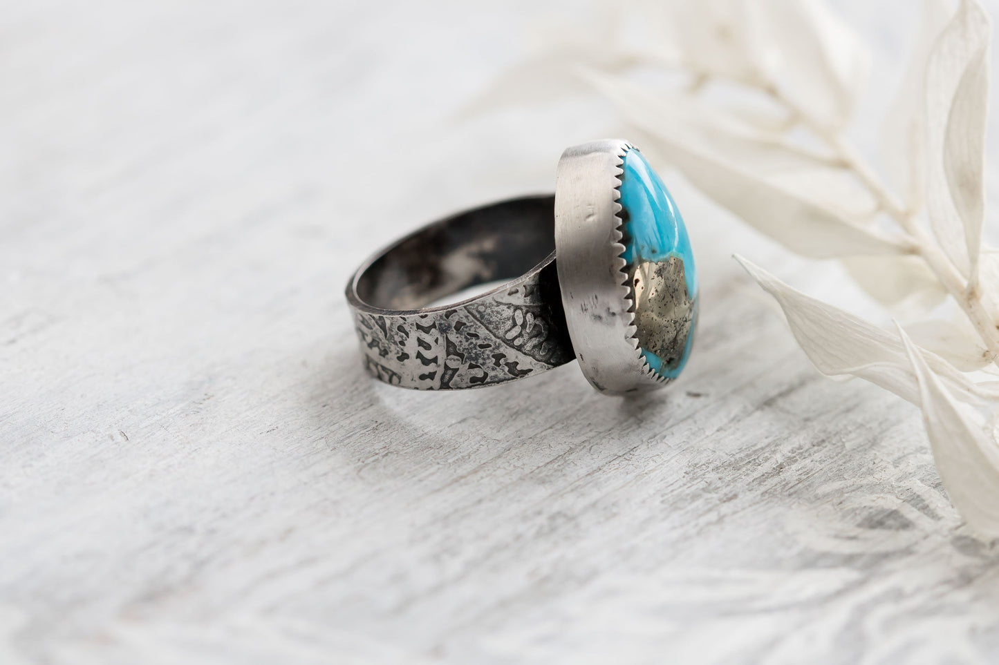 A sterling silver ring with a hand-cut Campitos turquoise stone featuring pyrite speckles, placed on a white wooden surface with dried white leaves in the background.