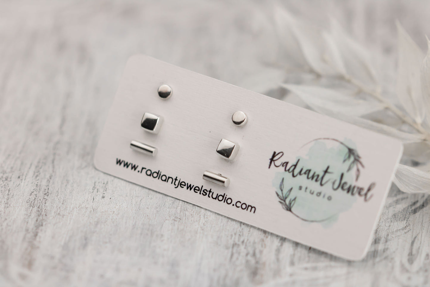 A set of minimalist sterling silver stud earrings presented on a white card with the Radiant Jewel Studio logo, featuring three different shapes: a dot, a square, and a bar.