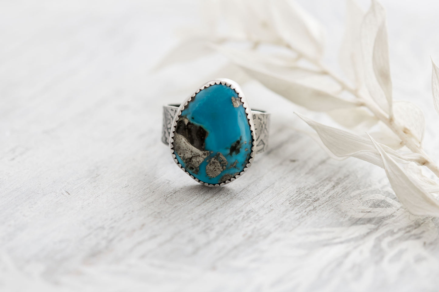 A sterling silver ring with a hand-cut Campitos turquoise stone featuring pyrite speckles, placed on a white wooden surface with dried white leaves in the background.