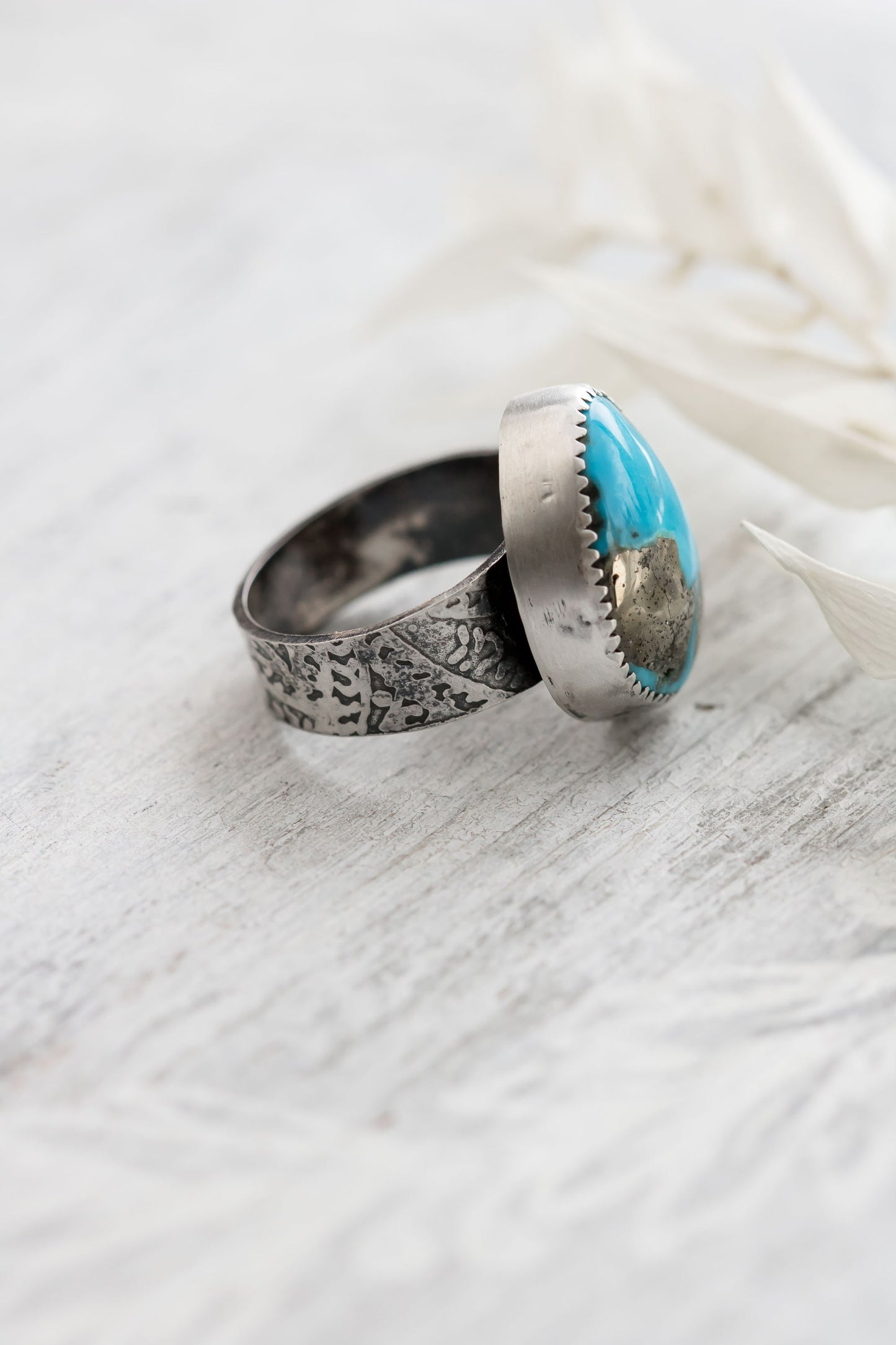 A sterling silver ring with a hand-cut Campitos turquoise stone featuring pyrite speckles, placed on a white wooden surface with dried white leaves in the background.