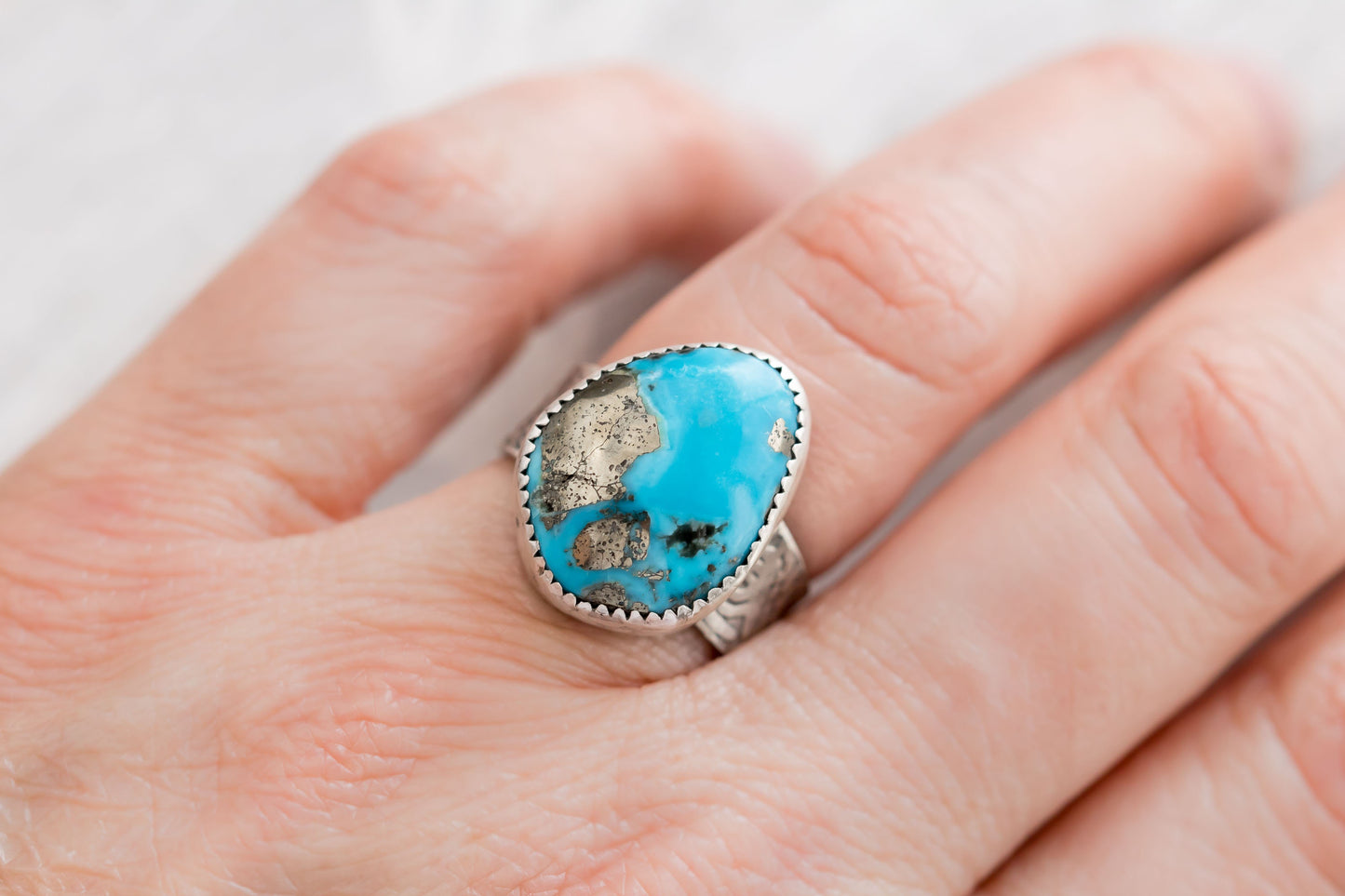 Close-up of a hand wearing a handcrafted sterling silver ring with a striking Campitos turquoise stone and pyrite inclusions, showcasing the textured silver band against a soft, blurred background.
