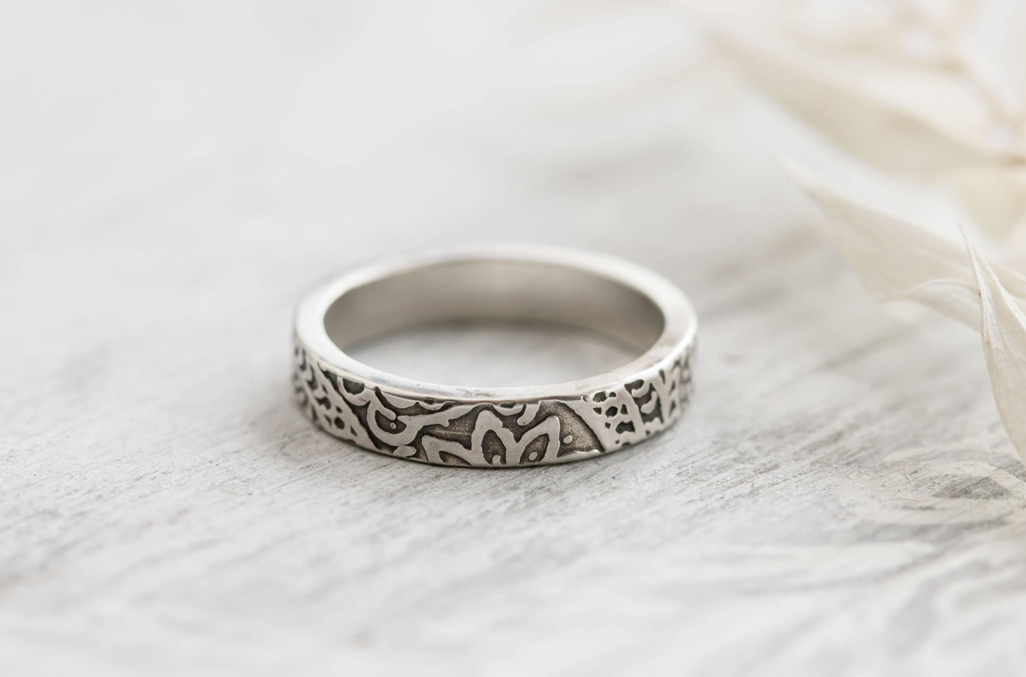 Detailed view of a sterling silver floral textured ring on a light wooden surface with soft floral accents, emphasizing the unique pattern.