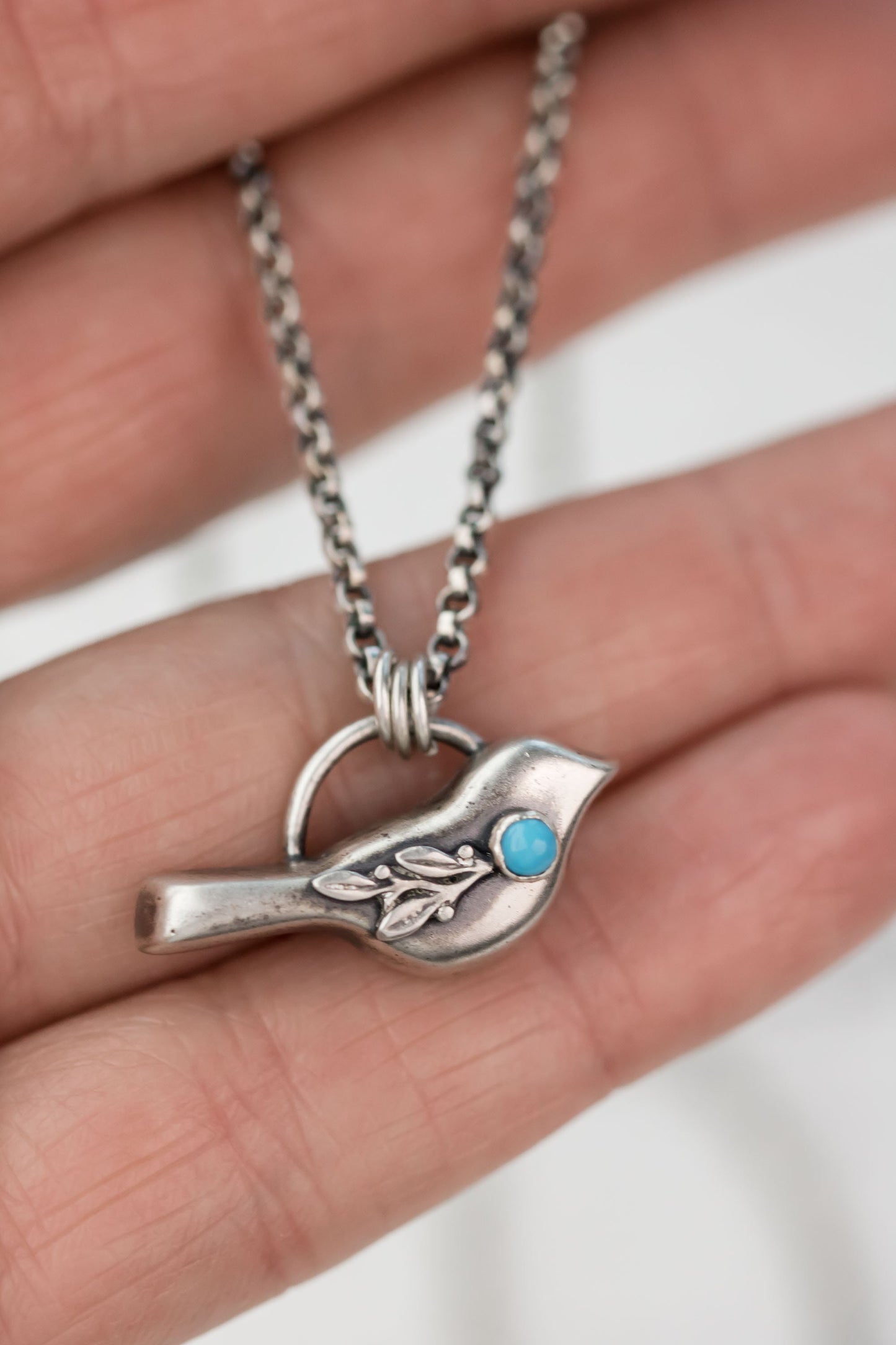 A hand holding a sterling silver bird necklace with a turquoise gemstone, showcasing the pendant’s floral details.