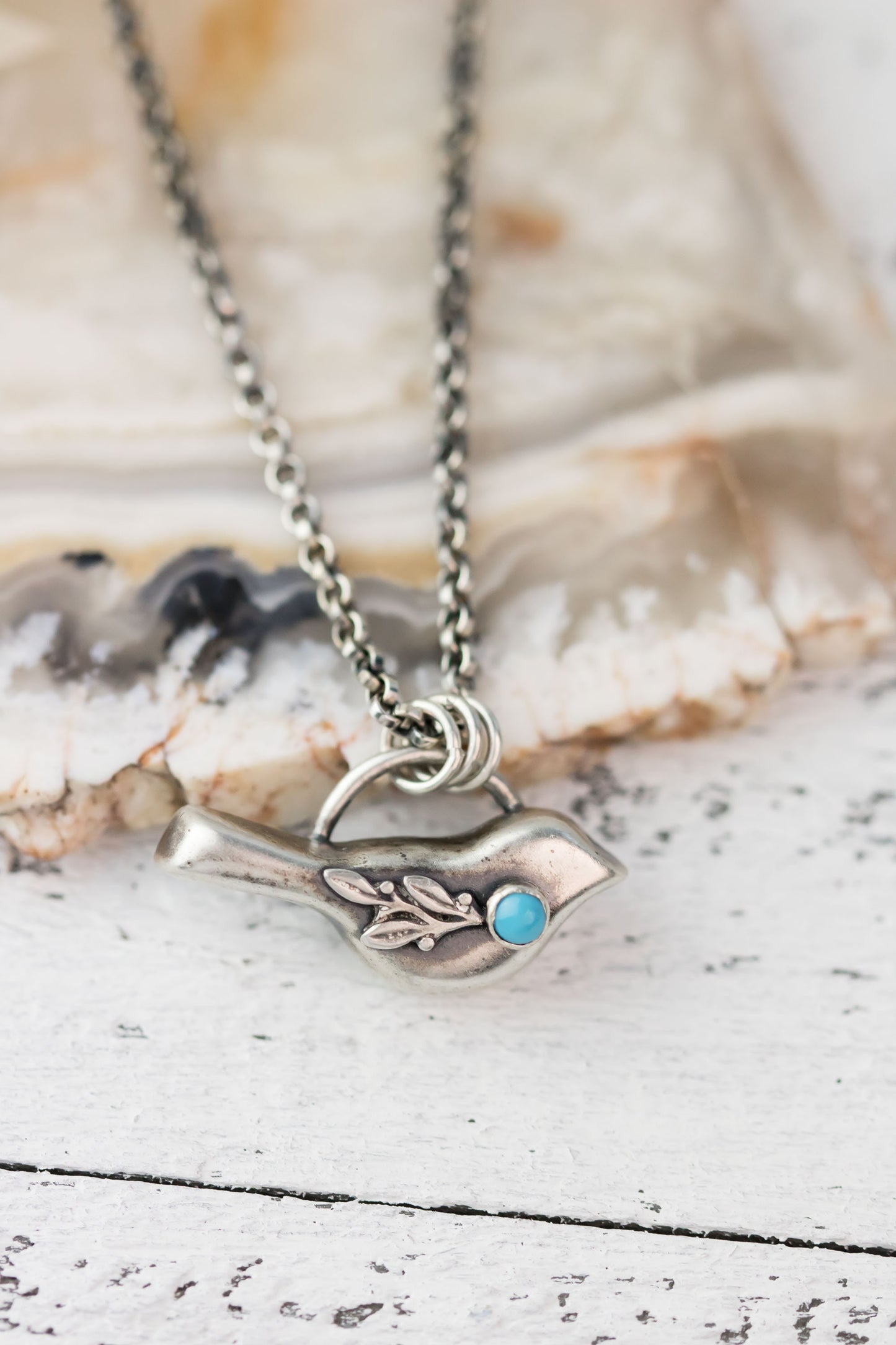 A sterling silver bird necklace with a turquoise gemstone accent, hanging on a rolo chain, displayed on a white wooden surface.