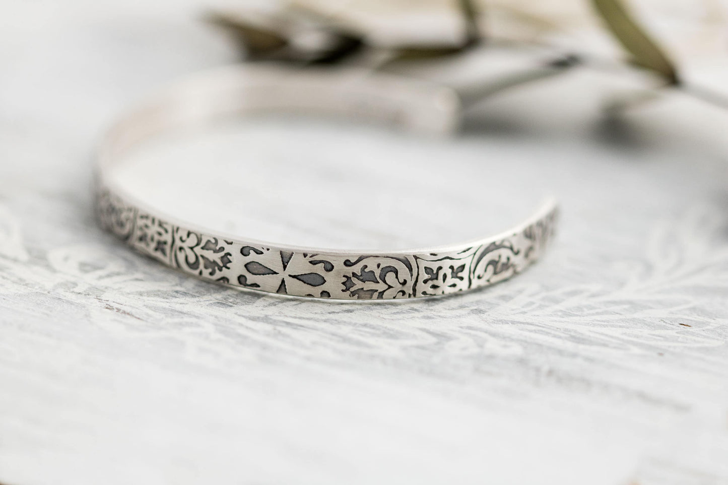A close-up photograph of a sterling silver cuff bracelet with intricate floral and scrollwork engravings. The bracelets have a slightly aged, oxidized finish that highlights the details of the patterns. They are displayed on a light, textured surface with soft-focus greenery and neutral-toned elements in the background.