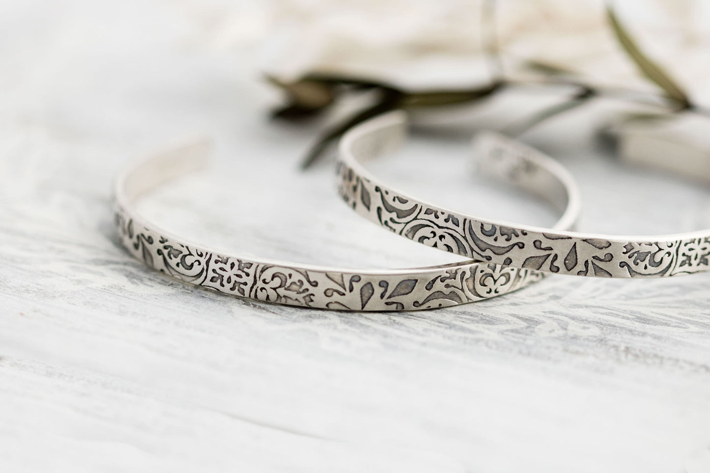A close-up photograph of two sterling silver cuff bracelets with intricate floral and scrollwork engravings. The bracelets have a slightly aged, oxidized finish that highlights the details of the patterns. They are displayed on a light, textured surface with soft-focus greenery and neutral-toned elements in the background.