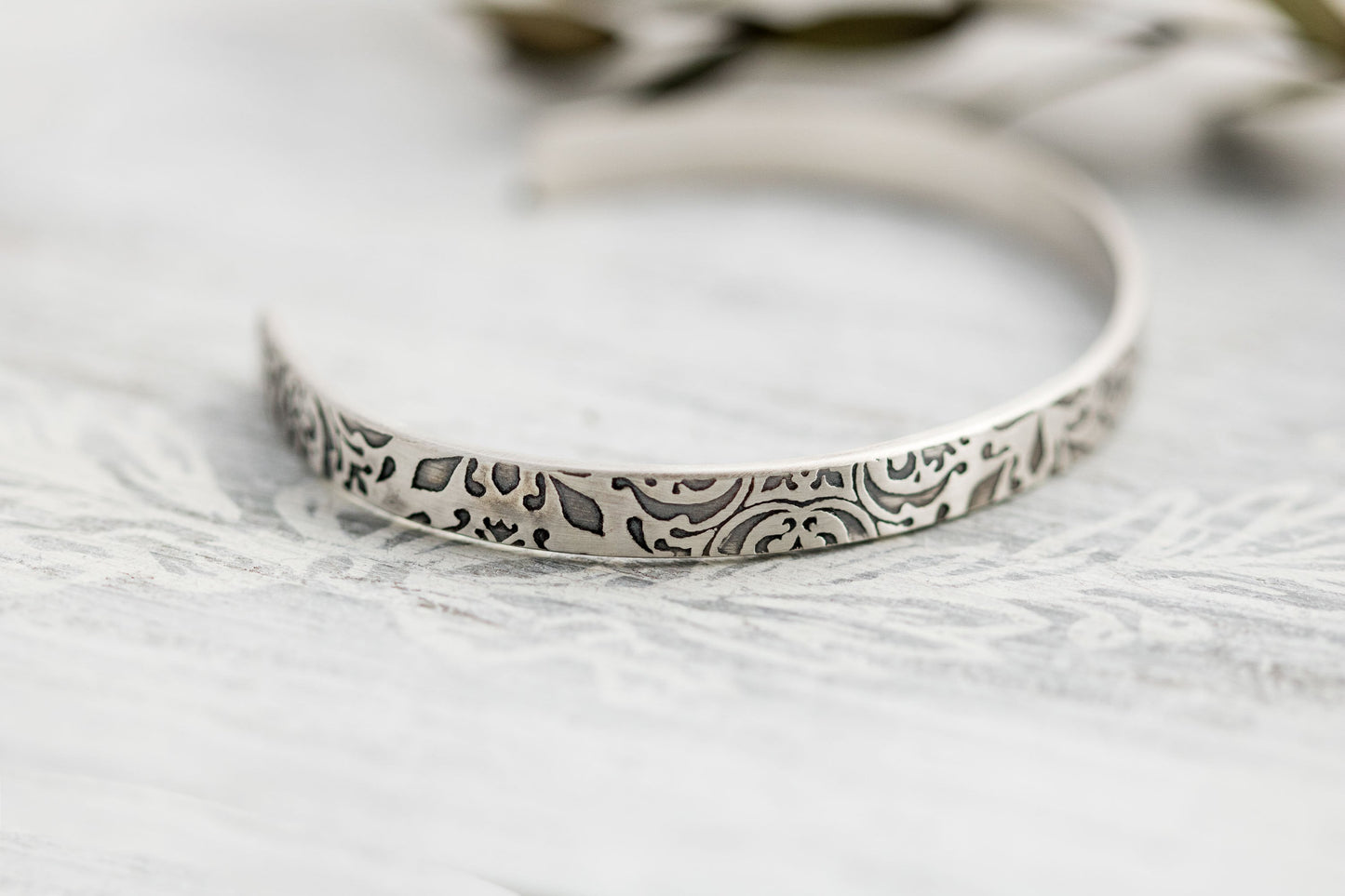 A close-up photograph of a sterling silver cuff bracelet with intricate floral and scrollwork engravings. The bracelets have a slightly aged, oxidized finish that highlights the details of the patterns. They are displayed on a light, textured surface with soft-focus greenery and neutral-toned elements in the background.