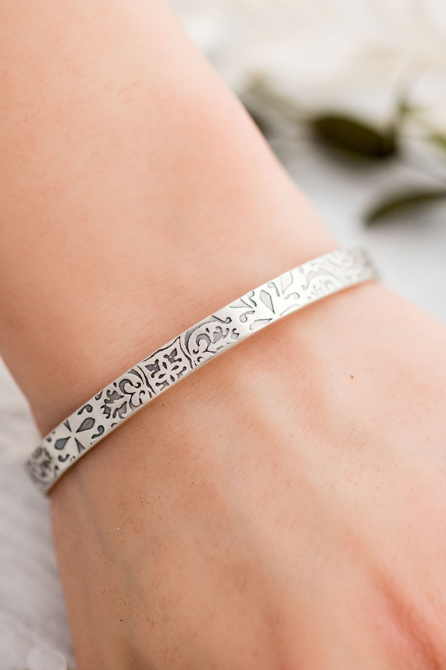 A close-up photograph of a sterling silver cuff bracelet on a wrist with intricate floral and scrollwork engravings. The bracelets have a slightly aged, oxidized finish that highlights the details of the patterns. They are displayed on a light, textured surface with soft-focus greenery and neutral-toned elements in the background.