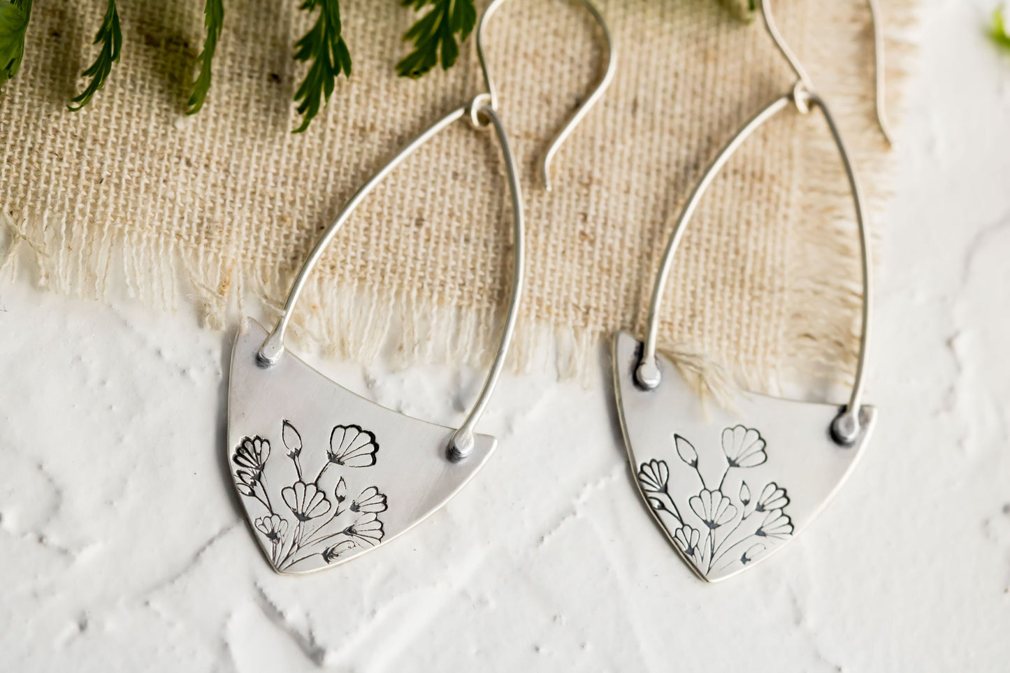 Handcrafted sterling silver Art Nouveau floral hoop earrings on a textured background with greenery.