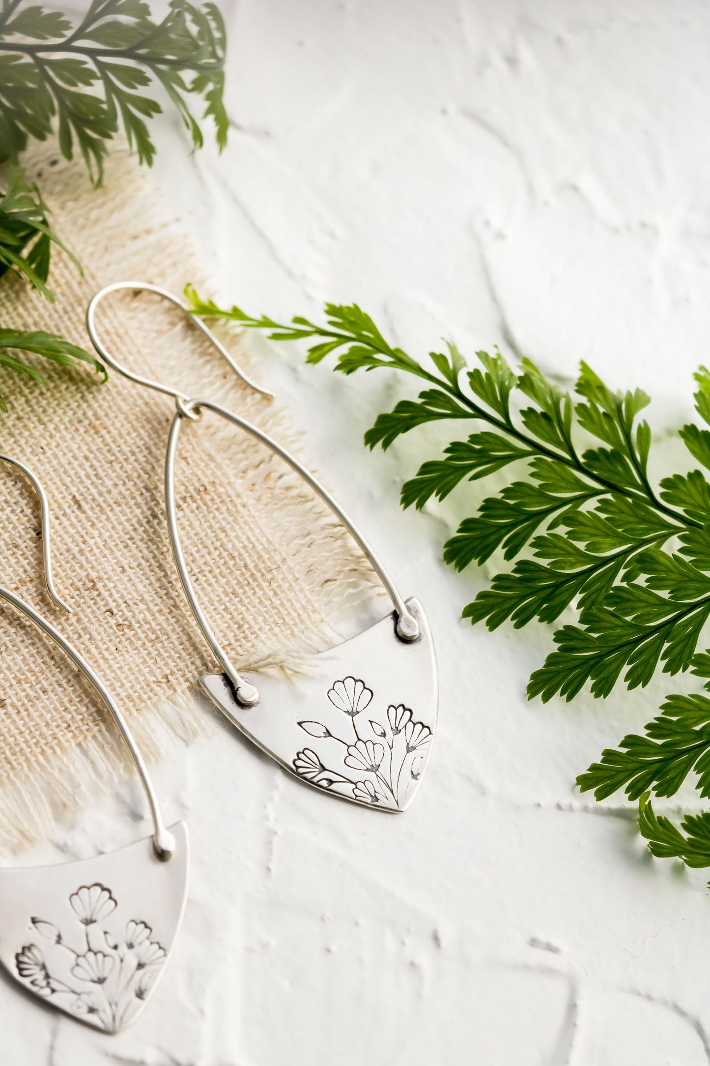 Handcrafted sterling silver Art Nouveau floral hoop earrings on a textured background with greenery.