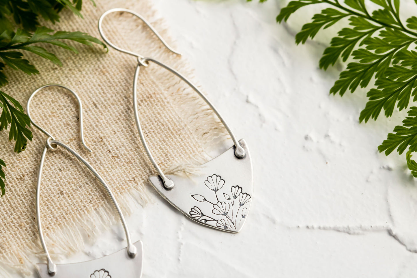 Handcrafted sterling silver Art Nouveau floral hoop earrings on a textured background with greenery.