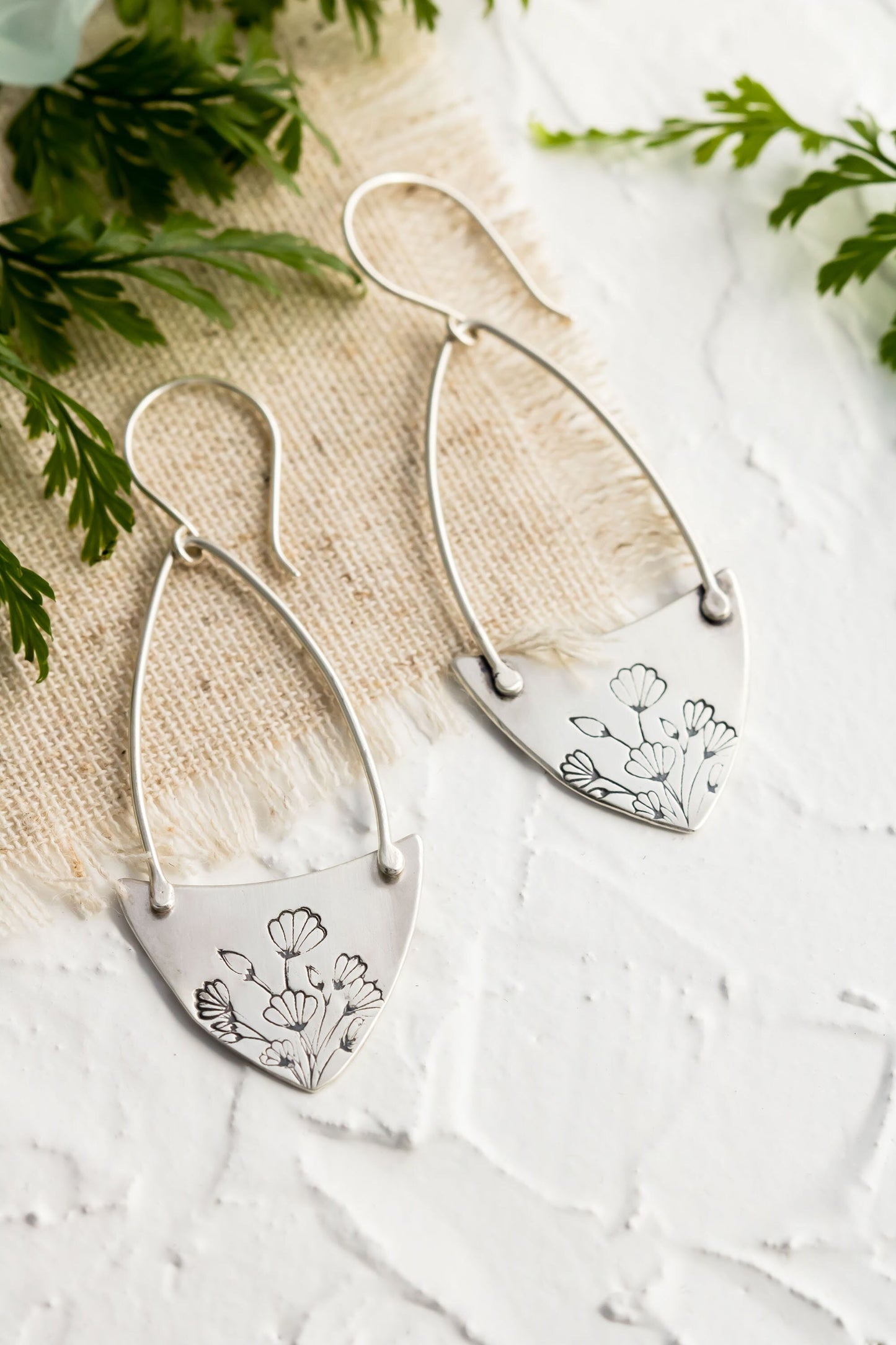 Handcrafted sterling silver Art Nouveau floral hoop earrings on a textured background with greenery.