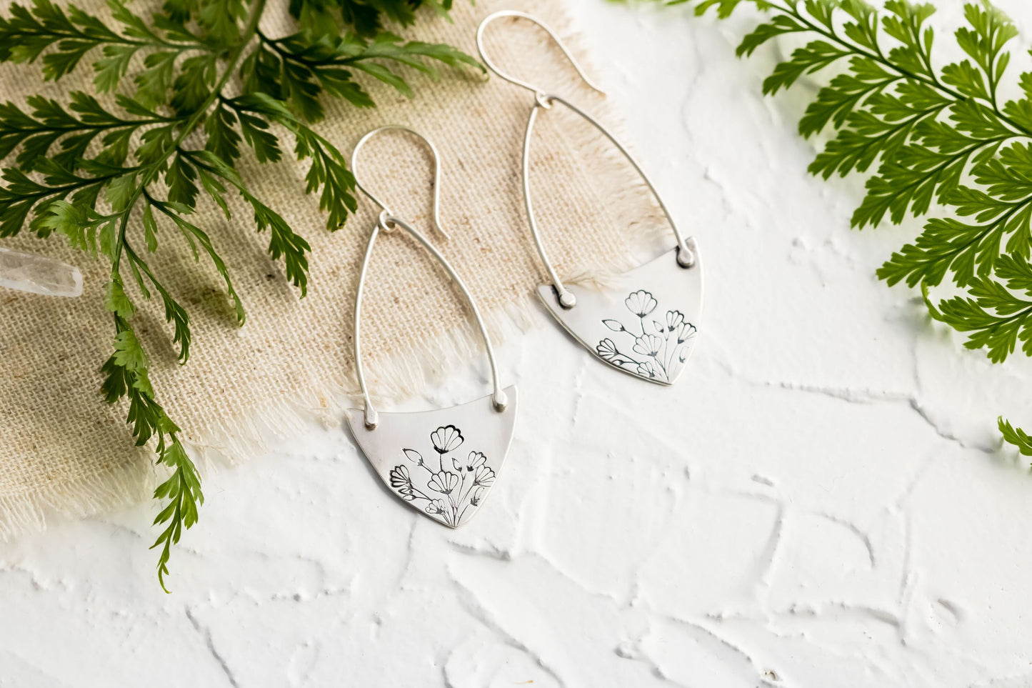 Handcrafted sterling silver Art Nouveau floral hoop earrings on a textured background with greenery.